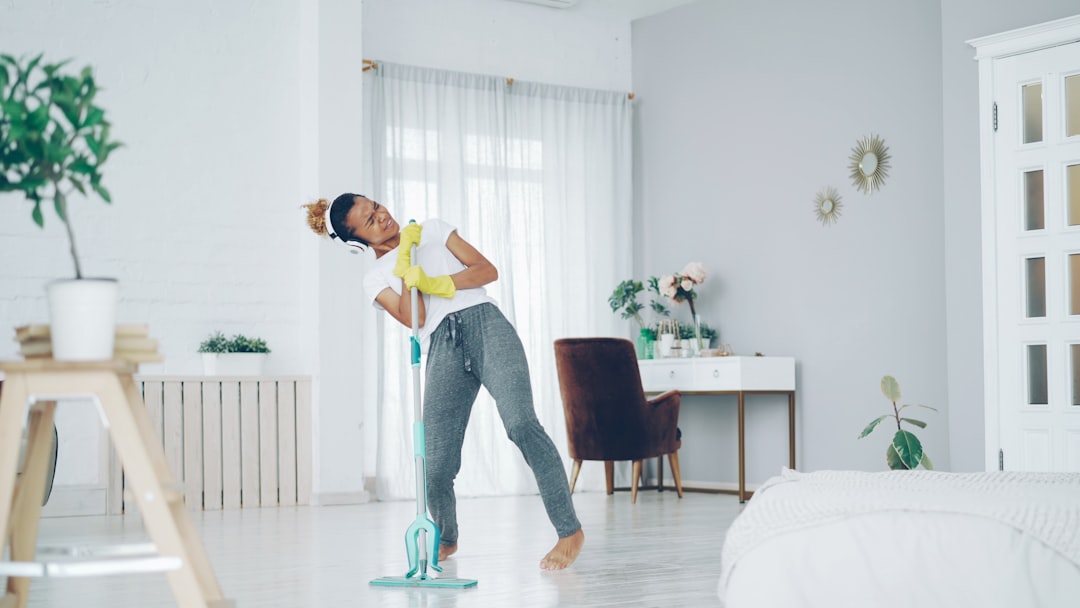Happy African American woman is washing floor with plastic mop, singing and dancing listening to music through headphones. People and modern technology concept.